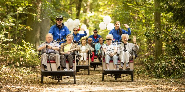 Ältere Menschen sitzen in Fahrrad-Rikschas und werden von anderen Personen durch einnen Wald kutschiert.