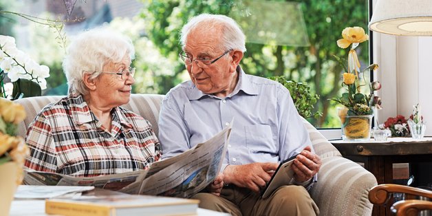©Ingo Bartussek_adobe.stock.com Alter Mann und alte Frau sitzen auf dem Sofa. Sie liest Zeitung, er schaut in ein Tablet.