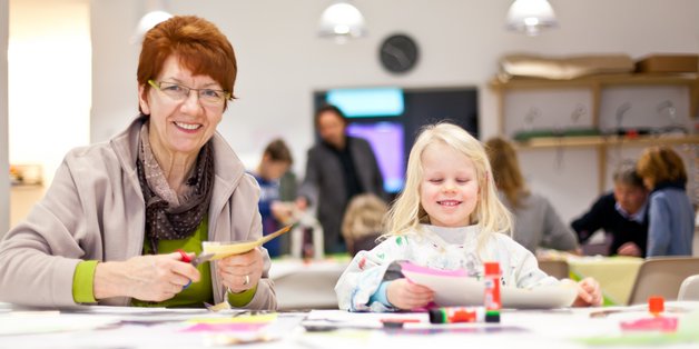 ©kristall_stock.adobe.com alte Frau und Kind basteln in einem Raum, in dem auch weitere Menschen sind.