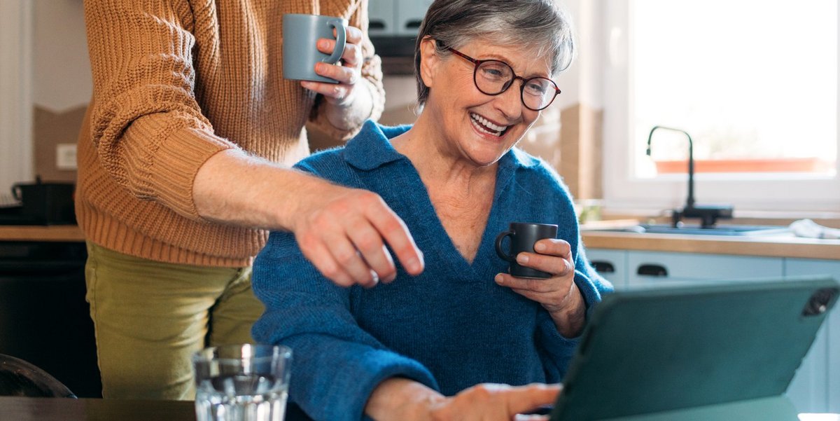 ©Studio Firma_stocky.com Eine ältere Frau mit kurzem grauen Haar, Brille und blauem Pullover sitzt am Küchentisch. Über ihr steht ein junger Mann mit Rollkragenpullower. Sie haben beide eine Tasse in der Hand und schauen auf ein Tablet, dass auf dem Tisch steht. Beide zeigen auf den Bildschirm.